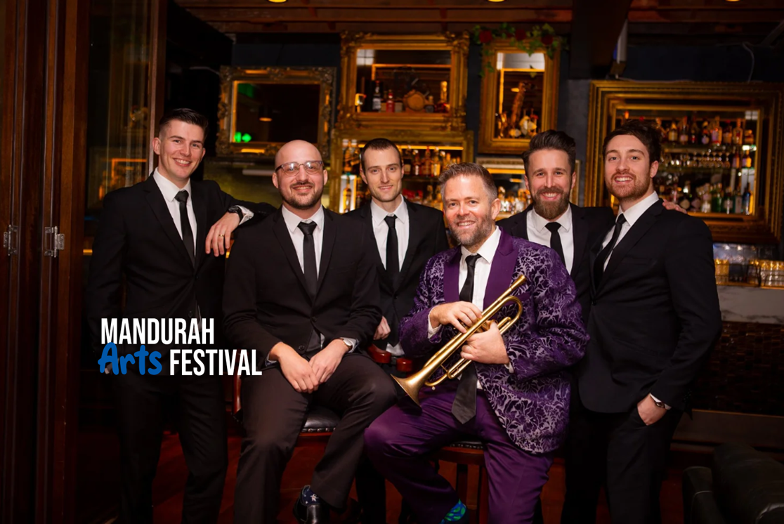 A group of men in velvet suits pose in front of a bar. One man's suit is purple and he holds a trumpet